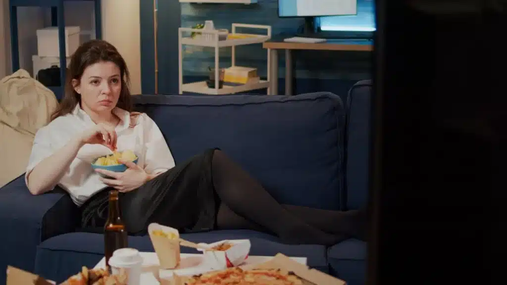 O papel da tecnologia na amplificação do problema da solidão Mulher assistindo televisão com um semblante triste, rodeada de comida, refletindo sobre como a tecnologia pode agravar sentimentos de solidão.