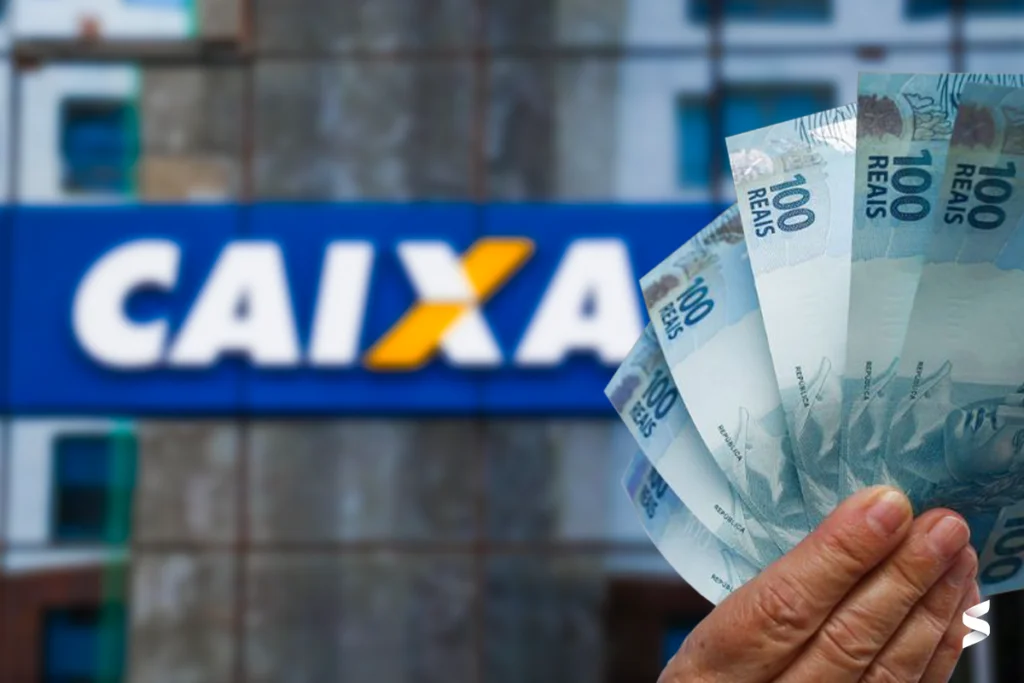 Mão segurando notas de R$ 100 em frente a uma agência da Caixa Econômica Federal.