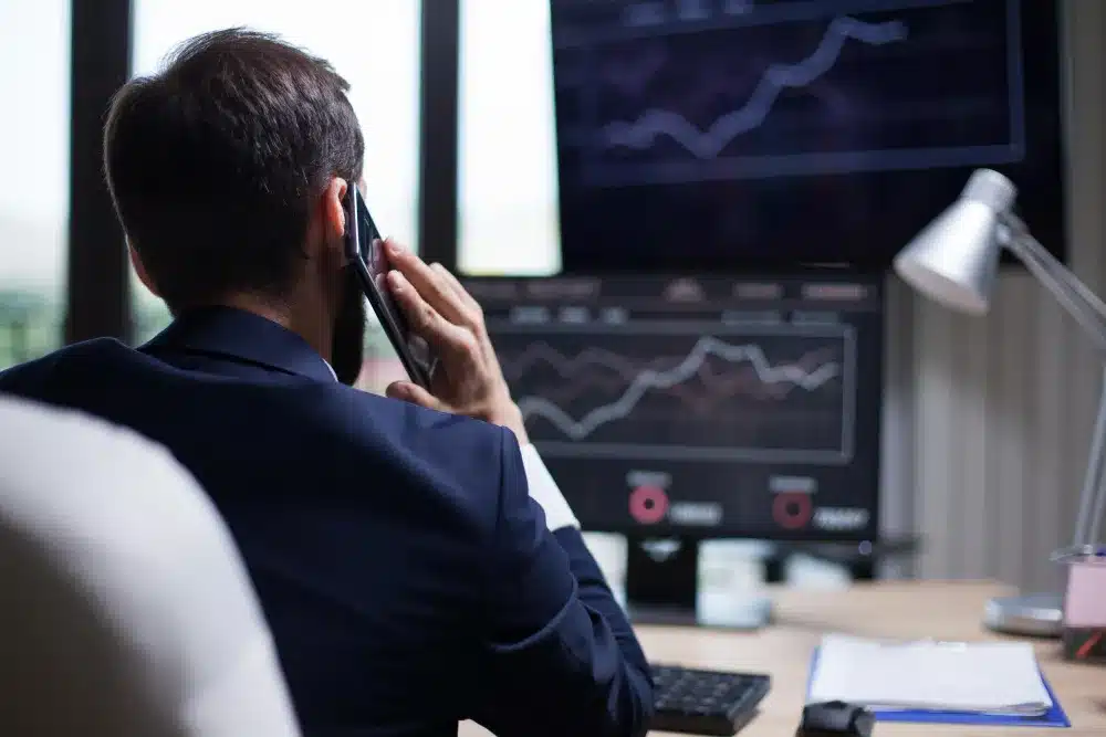 Trader analisando o mercado financeiro e negociando ativos. Trader observando gráficos financeiros em múltiplas telas enquanto fala ao telefone.