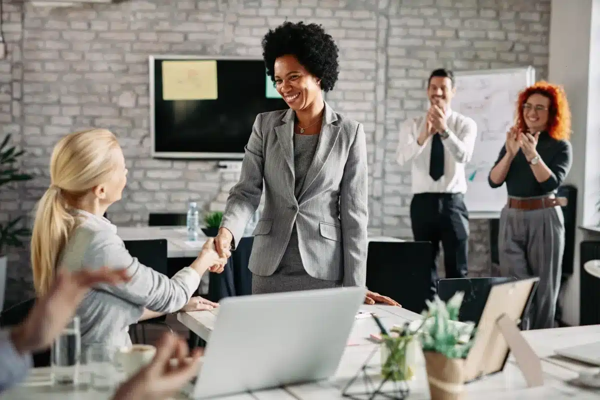 O Cursos de Inglês favorece a Valorização no Mercado de Trabalho em 2025 Profissionais comemorando a valorização no mercado de trabalho, com uma mulher recebendo aplausos e um aperto de mão.
