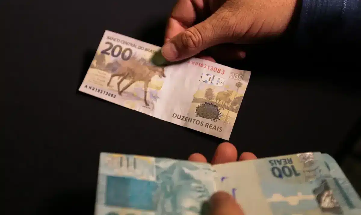 Pagamento do fundo PIS/Pasep sendo realizado com notas de R$ 100 e R$ 200. Pessoa segurando uma nota de R$ 200, enquanto outra nota de R$ 100 está visível na imagem, representando o valor disponível para saque.