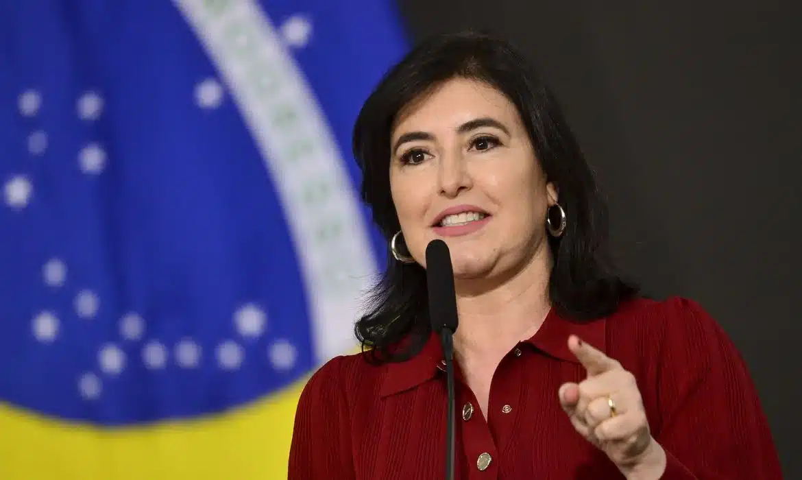O que foi anunciado para 2025 Imagem de uma mulher em evento oficial, com fundo da bandeira do Brasil, representando o que foi anunciado para 2025.