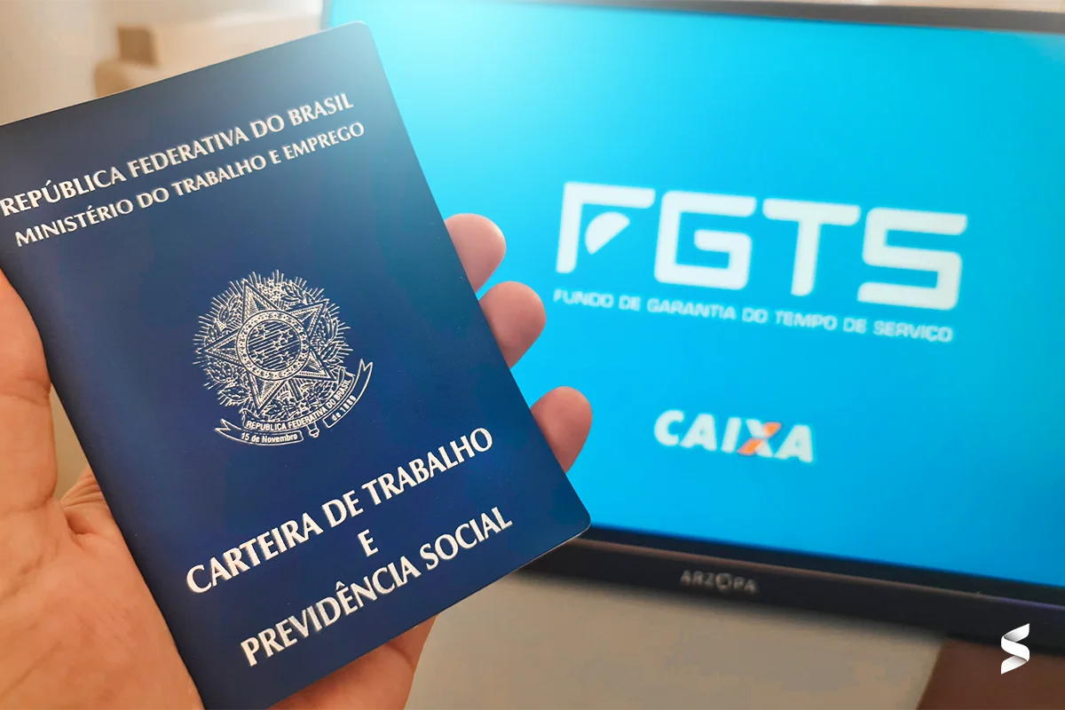 Saiba quem pode solicitar o saque-aniversário do FGTS e como fazer Pessoa segurando a carteira de trabalho enquanto consulta o FGTS no computador.