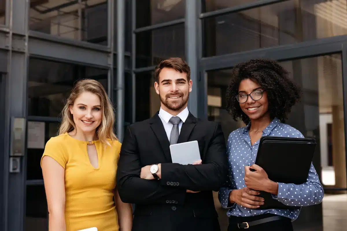 Três profissionais sorrindo para a foto em frente a um prédio corporativo, com diferentes dispositivos em mãos.