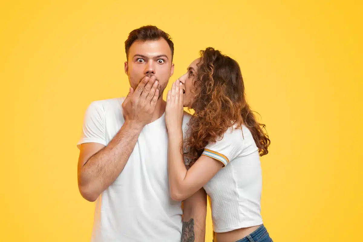 Casal sussurrando segredo, mulher falando no ouvido do homem com expressão surpresa, em um fundo amarelo vibrante.