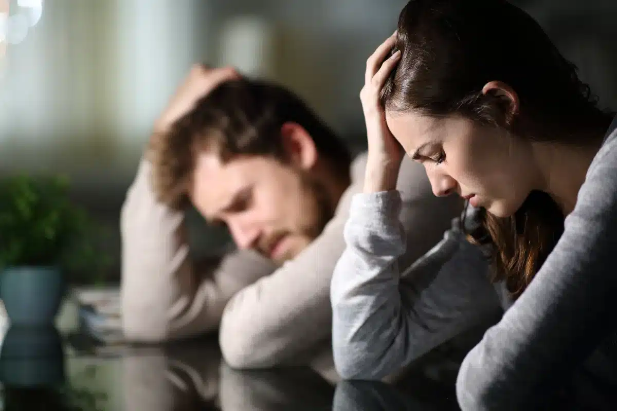 Casal passando por dificuldades emocionais Casal em conflito, com ambos visivelmente estressados e segurando a cabeça enquanto estão sentados à mesa.