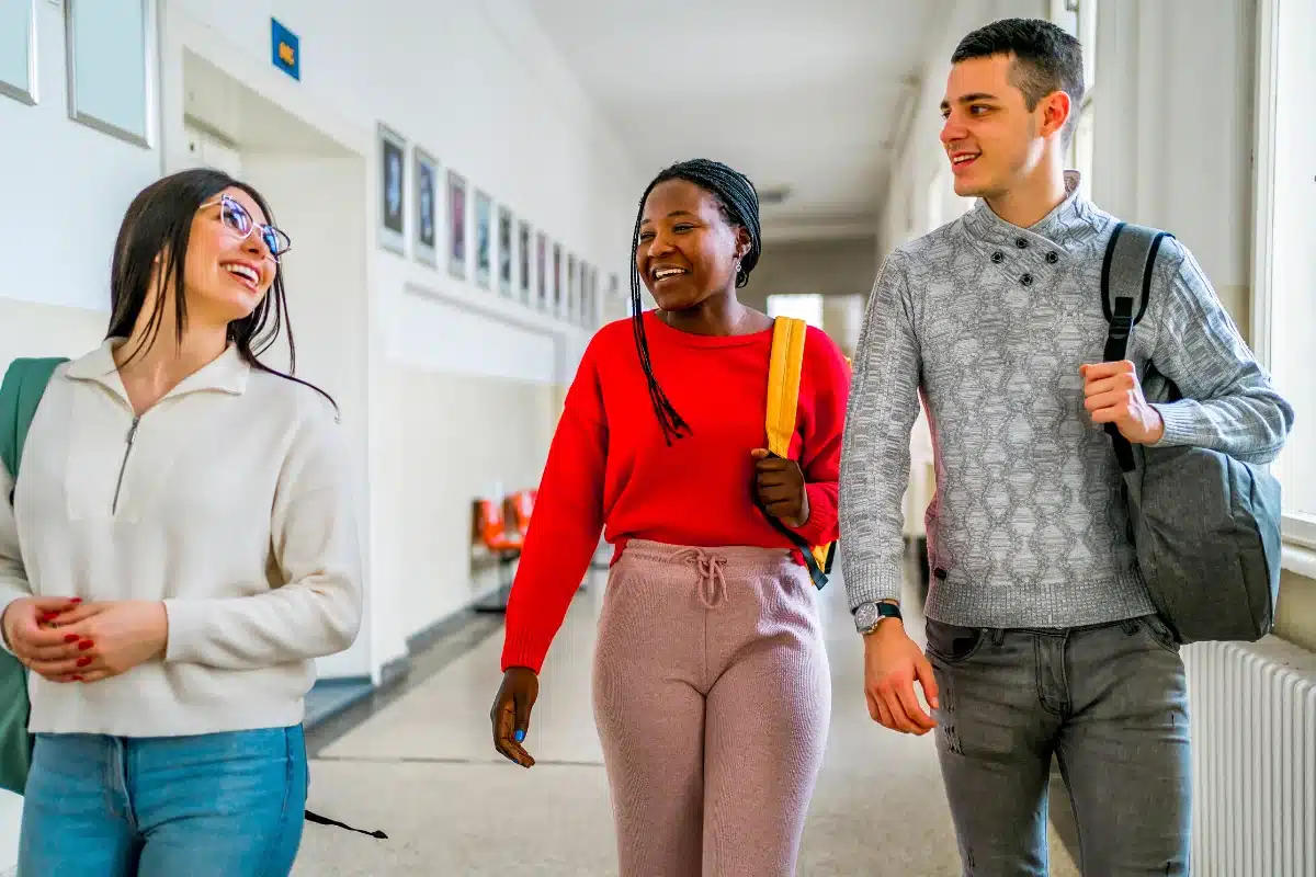 Informações Prouni Três estudantes sorrindo e caminhando no corredor da escola, com mochilas, felizes e interagindo.