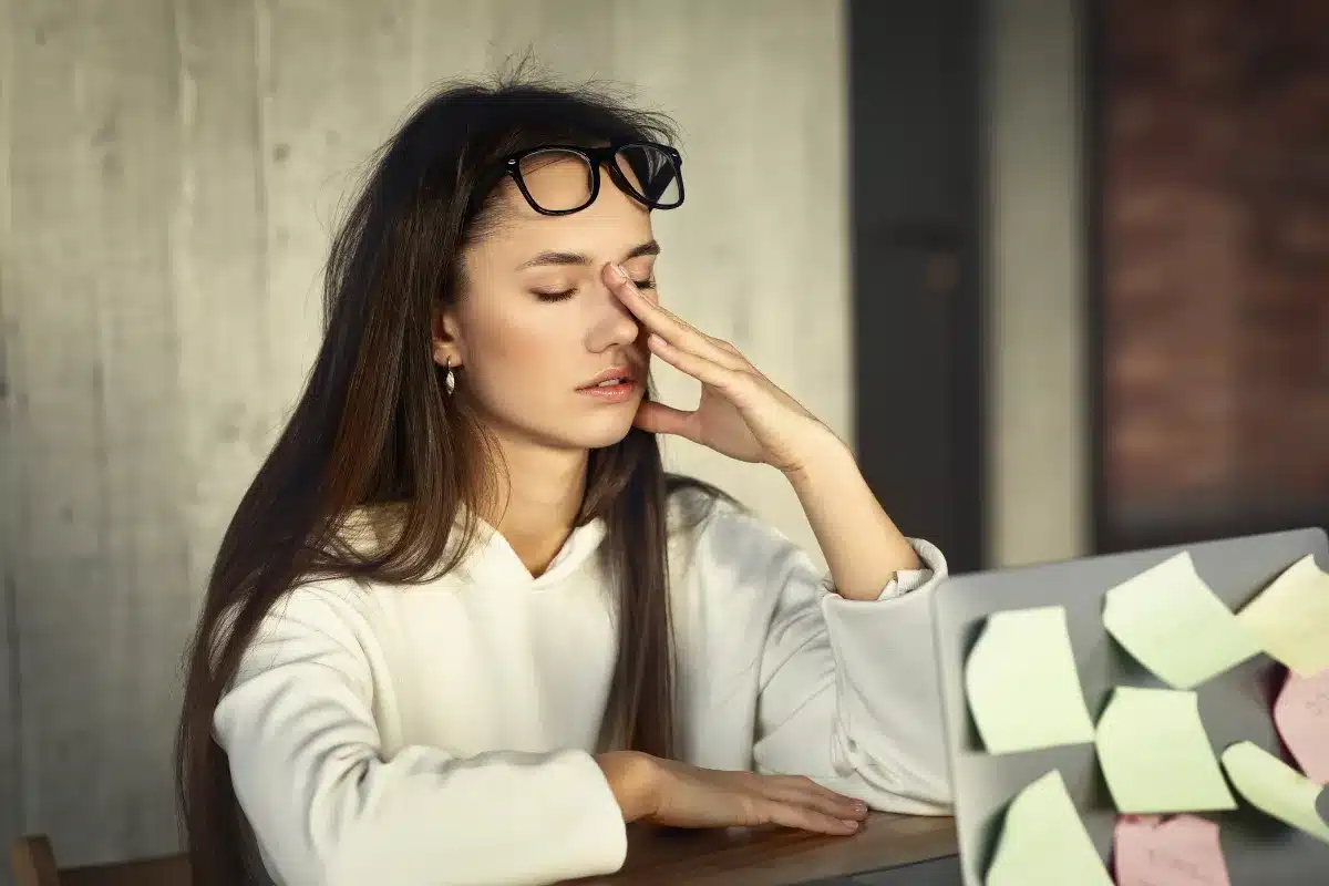 Mulher com expressão de cansaço e estresse enquanto trabalha, com óculos na cabeça e mãos no rosto, rodeada de post-its coloridos.
