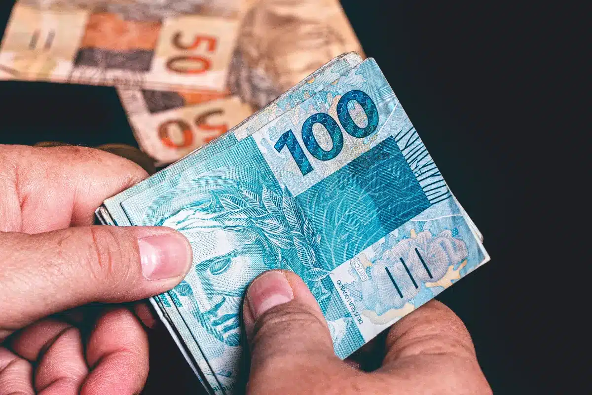 Pessoa contando cédulas de dinheiro em reais, com destaque para nota de R$ 100.