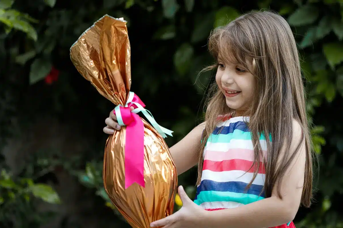 Menina com ovo de chocolate dourado Menina sorrindo ao segurar um grande ovo de chocolate dourado com fitas coloridas em um fundo de plantas verdes.