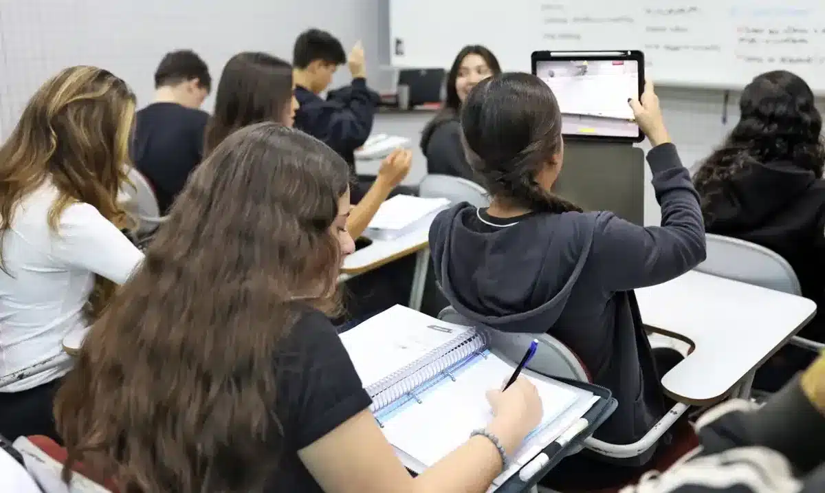 Estudantes em sala de aula utilizando cadernos e tablets durante atividade escolar.