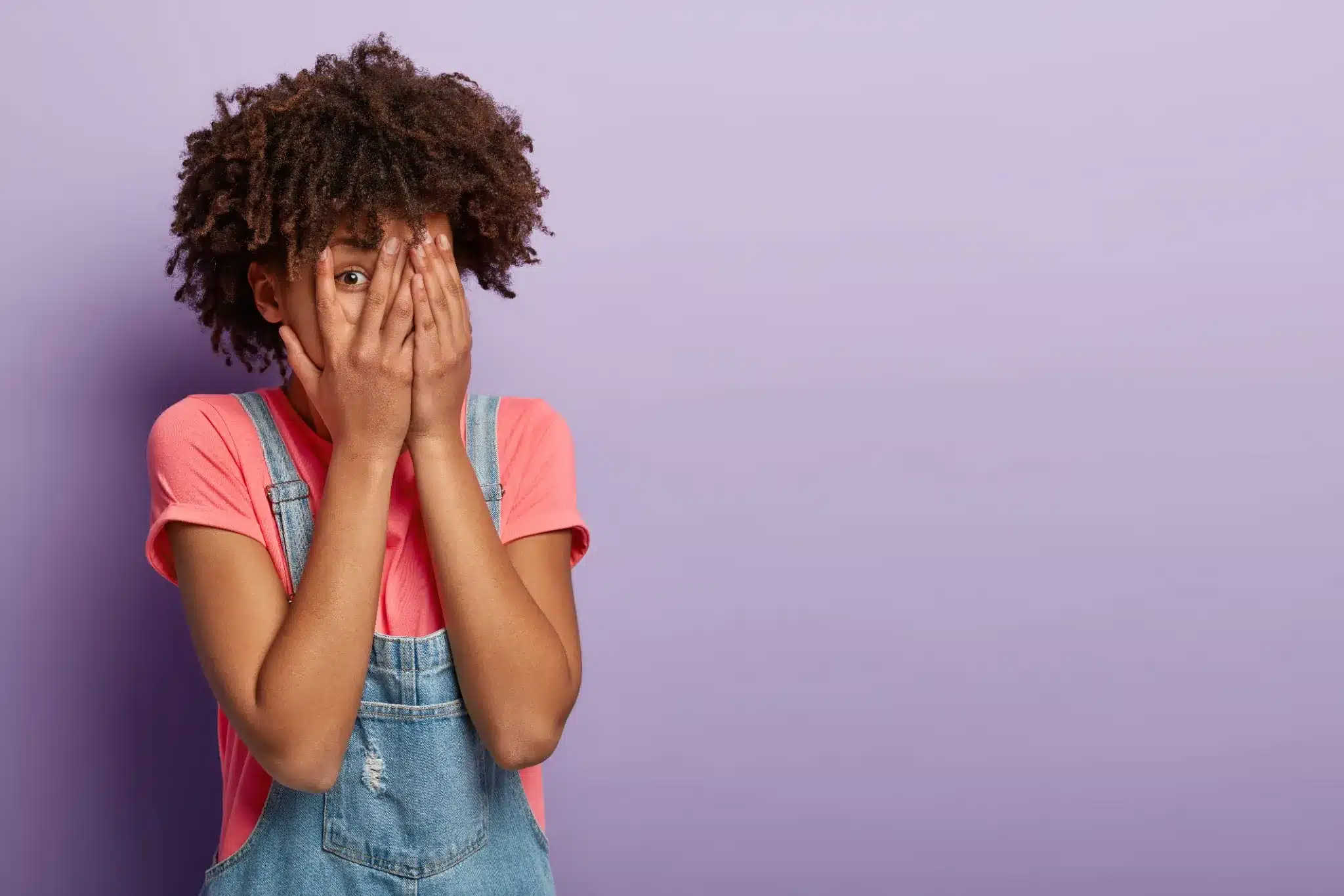 Jovem mulher com cabelo afro cobrindo parcialmente o rosto, expressando timidez em fundo roxo.