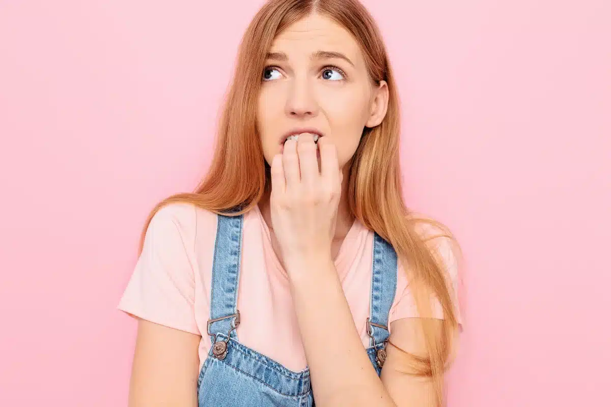 Mulher demonstrando insegurança, mordendo as unhas, em um fundo rosa.
