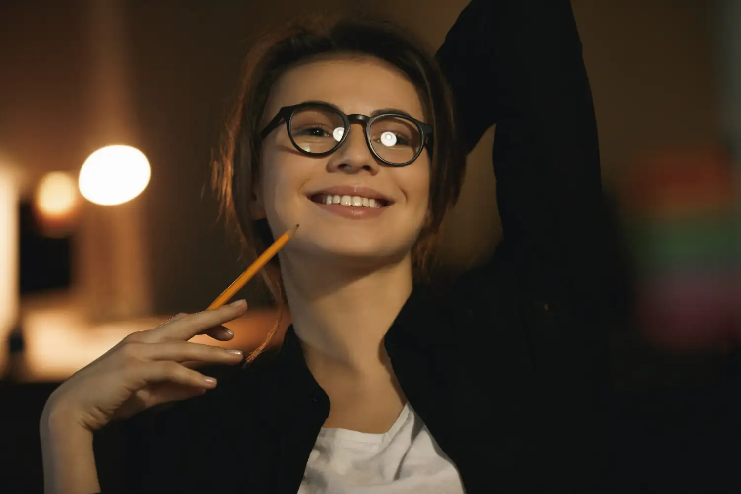 Jovem sorridente com aparência de concentração Jovem com óculos sorrindo enquanto segura um lápis, em ambiente de estudo.