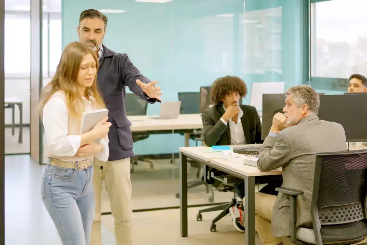 Humilhação no ambiente de trabalho: impacto no emocional Homem repreendendo mulher em ambiente de trabalho, com outros colegas observando a cena em um escritório moderno.
