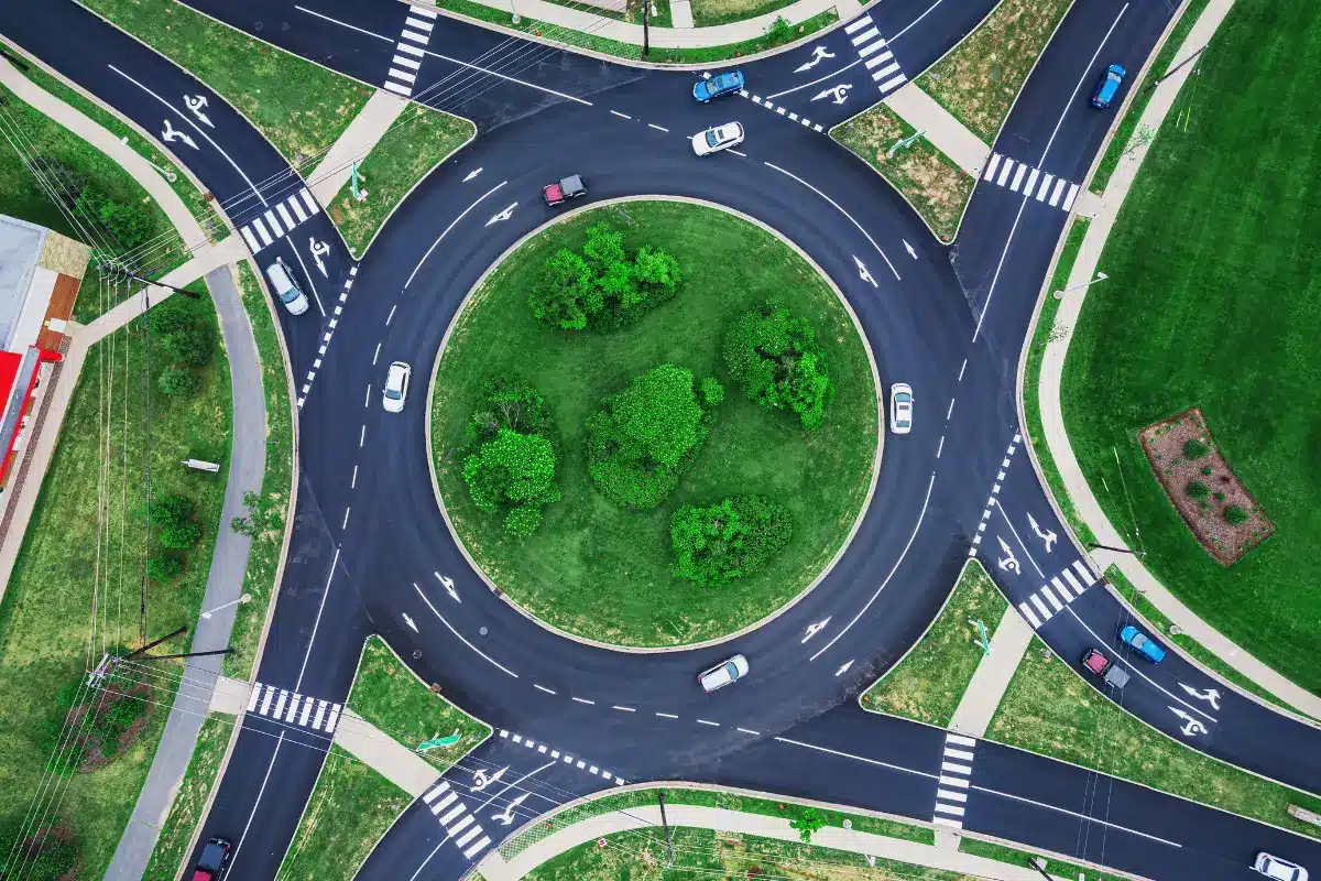Rotatória vista de cima com carros e vegetação ao redor Vista aérea de uma rotatória com carros circulando, cercada por calçadas e vegetação.