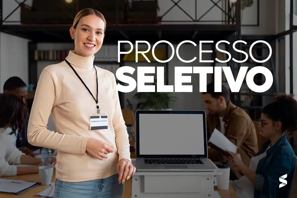 Profissional sorrindo em ambiente de trabalho, com computador à sua frente, representando o processo seletivo.