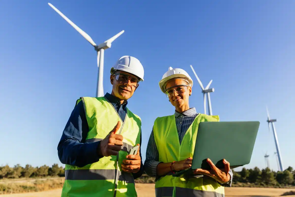 Profissionais em campo de energia renovável com turbinas eólicas Dois profissionais de energia renovável em um campo com turbinas eólicas, sorrindo enquanto operam dispositivos.