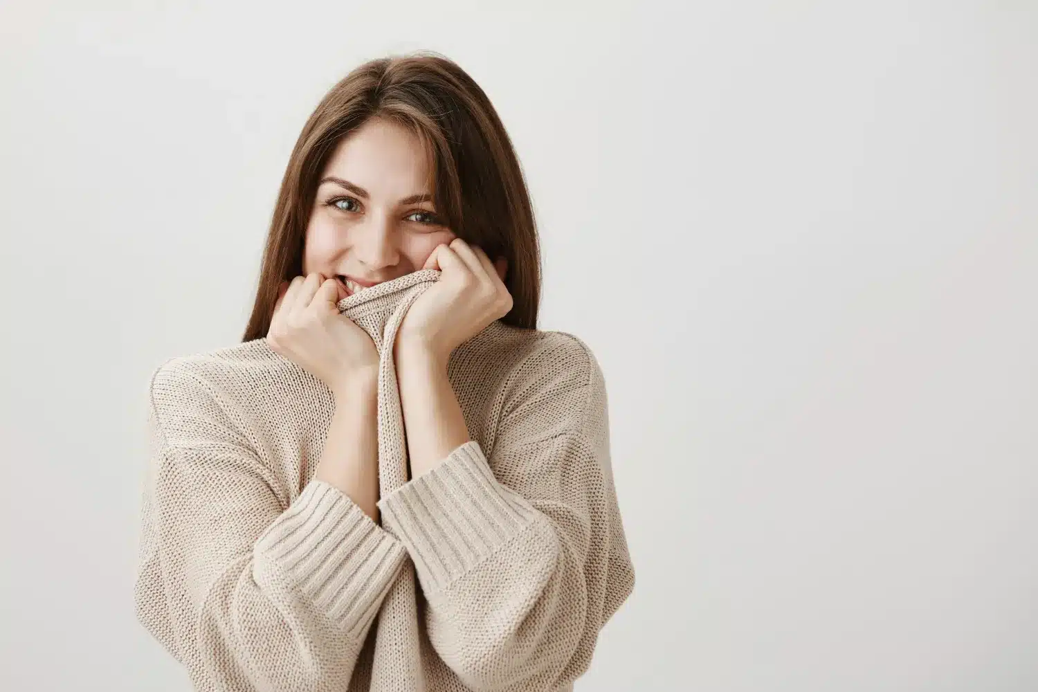 Mulher sorrindo timidamente, segurando a gola de um suéter bege, transmitindo a sensação de timidez e charme.