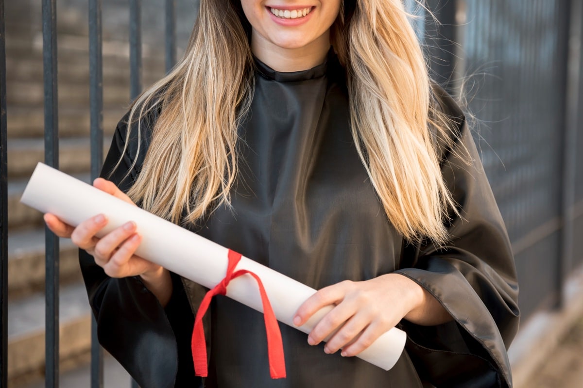 Jovem sorridente em beca segurando diploma com fita vermelha, simbolizando sucesso acadêmico e profissional.