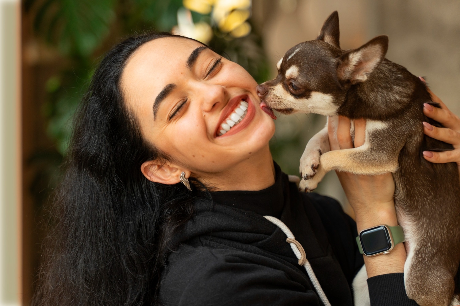 Mulher jovem segurando um cachorro pequeno que lambe seu rosto, ambos demonstrando afeto.