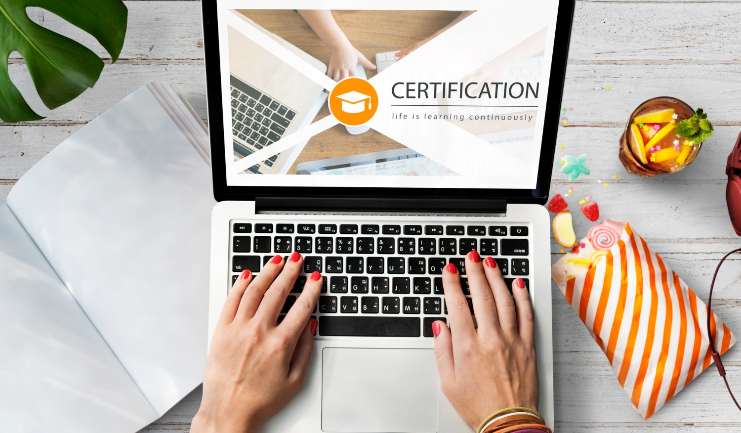 Pessoa digitando em notebook com tela mostrando palavra "CERTIFICATION" e símbolo de graduação, ao lado itens de estudo e lanche
