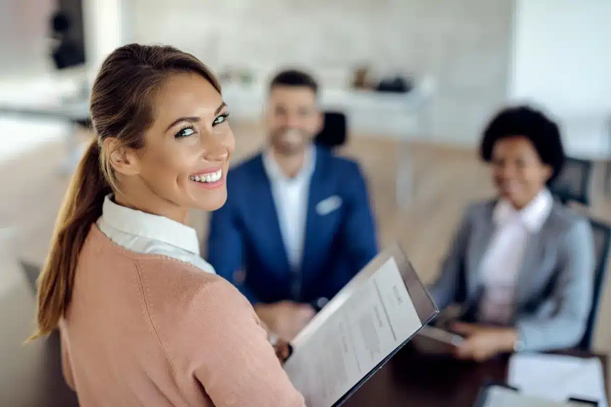 Feira de empregos e estágios: Programação de atividades para os participantes. Mulher sorrindo segurando seu currículo em frente a uma mesa com outros profissionais durante uma feira de empregos.