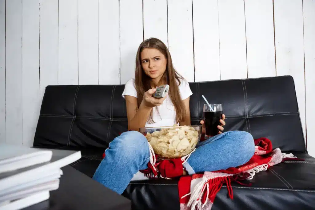 Como a comida favorita influencia a gestão do estresse Jovem assistindo TV com expressão pensativa, segurando um copo de refrigerante e uma tigela de batatas chips