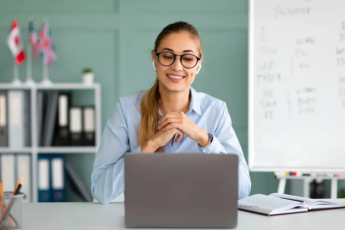 Ensino de línguas Mulher sorrindo, usando óculos e fones de ouvido, em frente a um laptop em uma sala de aula moderna.