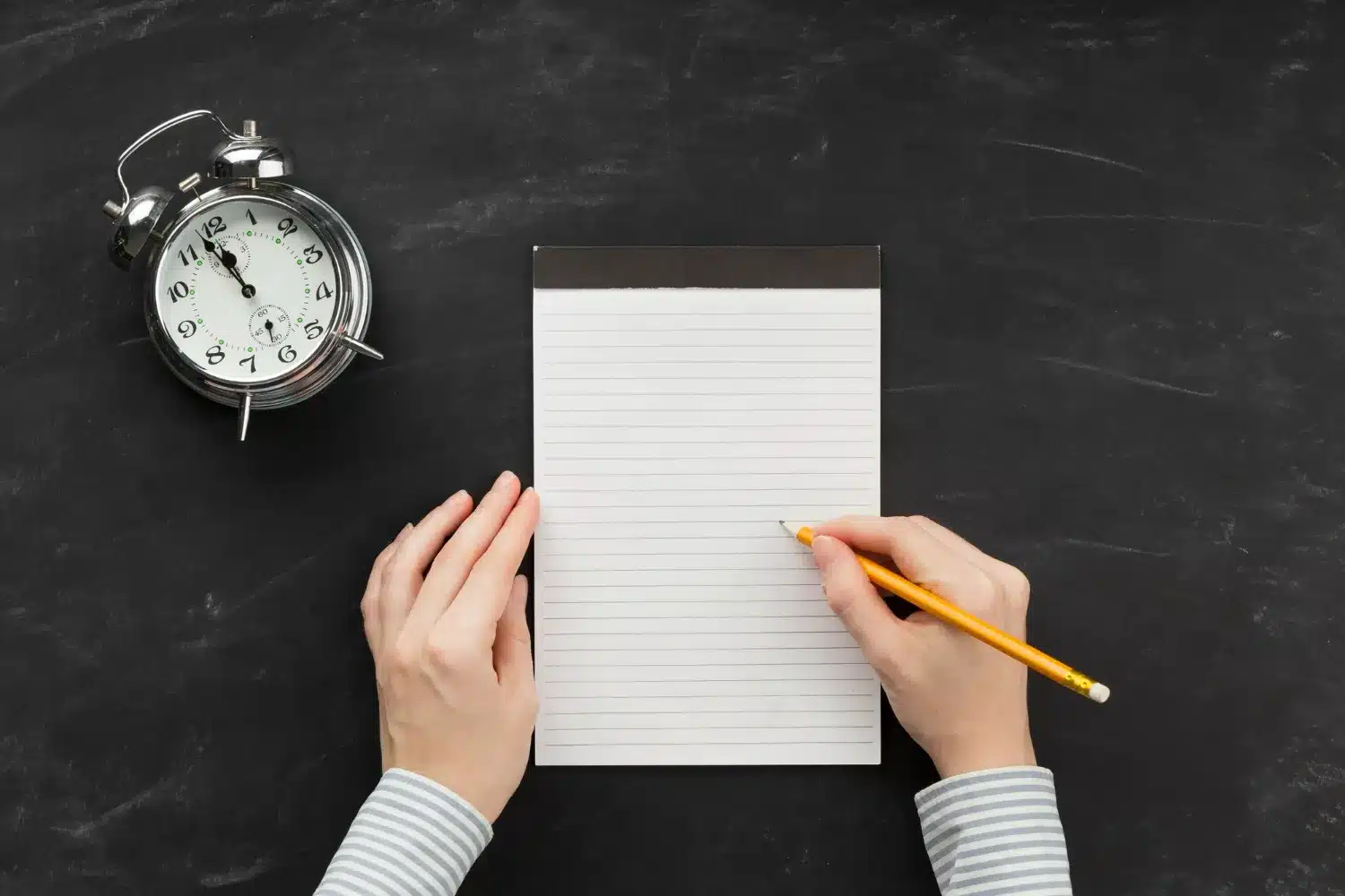 Bloco de notas e relógio analógico sobre mesa preta Mãos segurando lápis e bloco de notas ao lado de relógio analógico sobre mesa preta.
