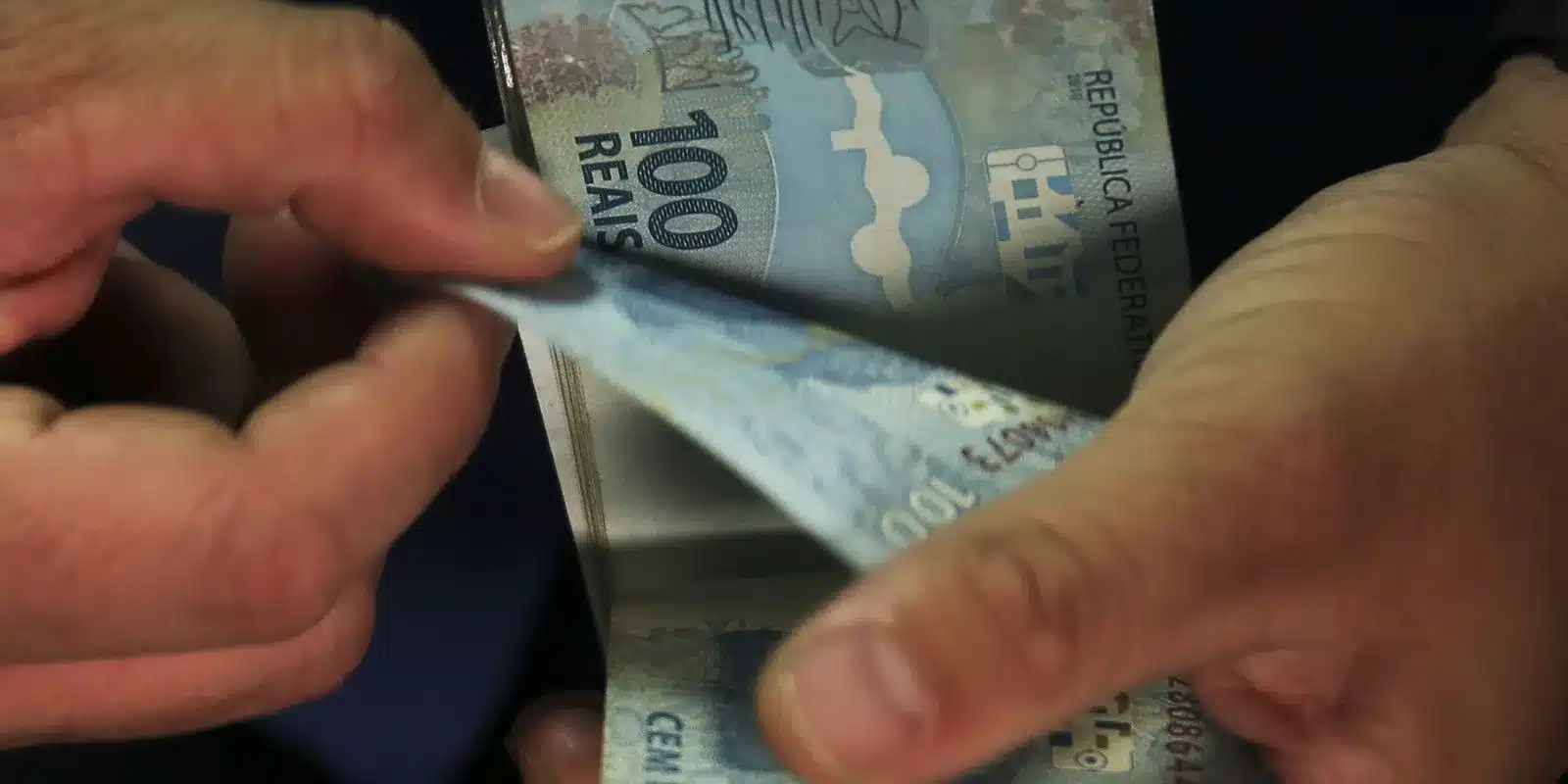 Pagamento da bolsa - Pé de Meia Licenciaturas Nota de 100 reais sendo manuseada por mãos, representando transação financeira.