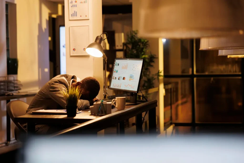Esgotamento mental no ambiente de trabalho Homem exausto no escritório com a cabeça abaixada sobre a mesa, simbolizando os efeitos do Burnout
