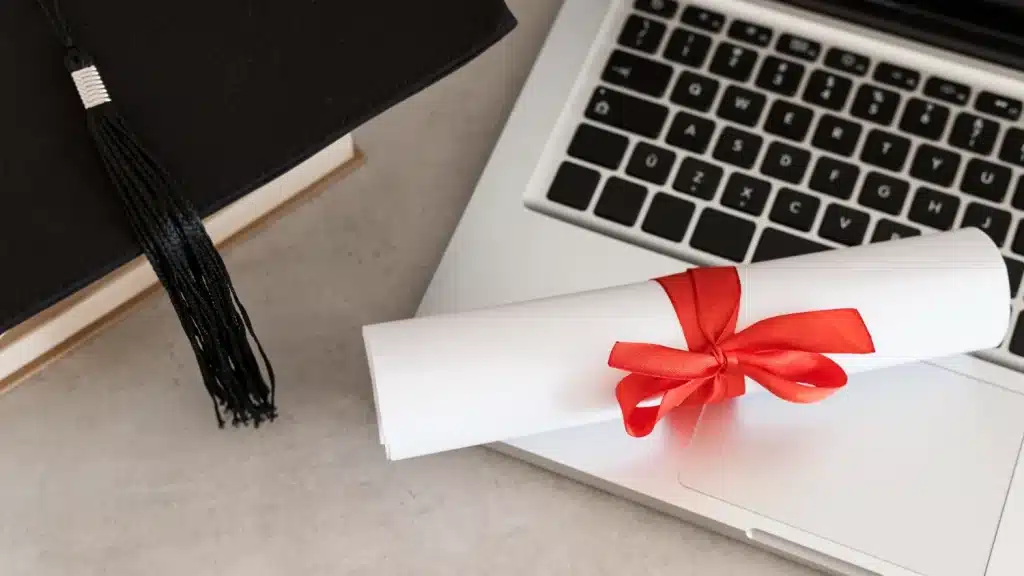 Certificação de curso online válida para concursos Certificado enrolado com laço vermelho sobre laptop e capelo, representando conclusão de curso online