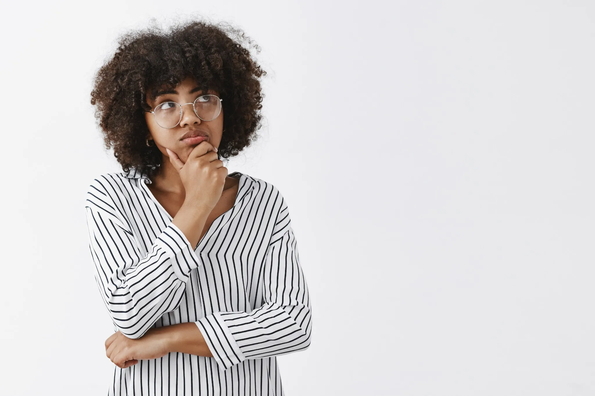 Mulher pensativa com cabelo cacheado, usando óculos e fazendo gesto de reflexão, com o rosto voltado para a direita.