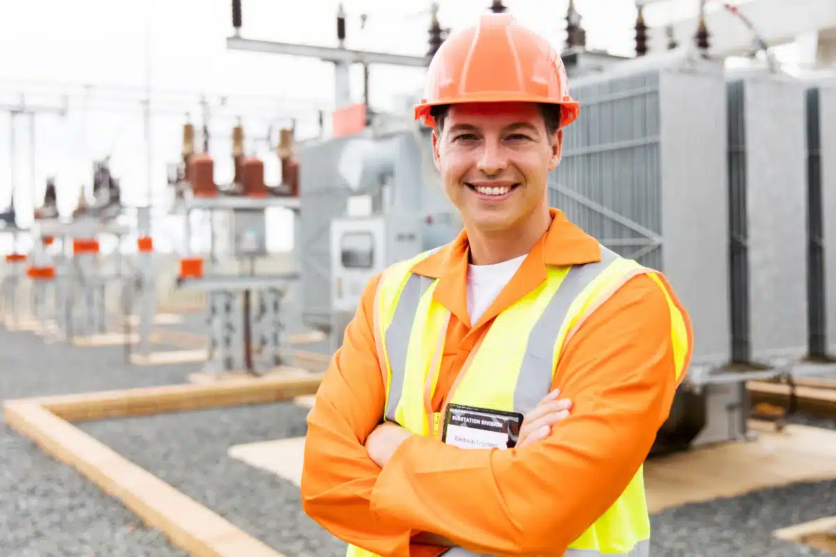 Engenheiro de segurança em campo, com uniforme de segurança e capacete, sorrindo em frente à estação de energia.