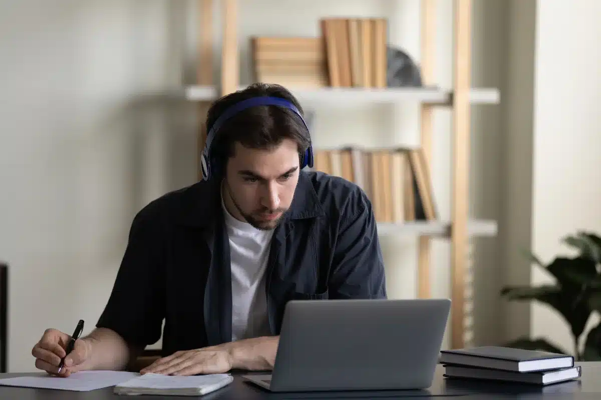 Estudante Aprendendo Espanhol Homem concentrado, usando fones de ouvido, estudando no computador em uma mesa com anotações.