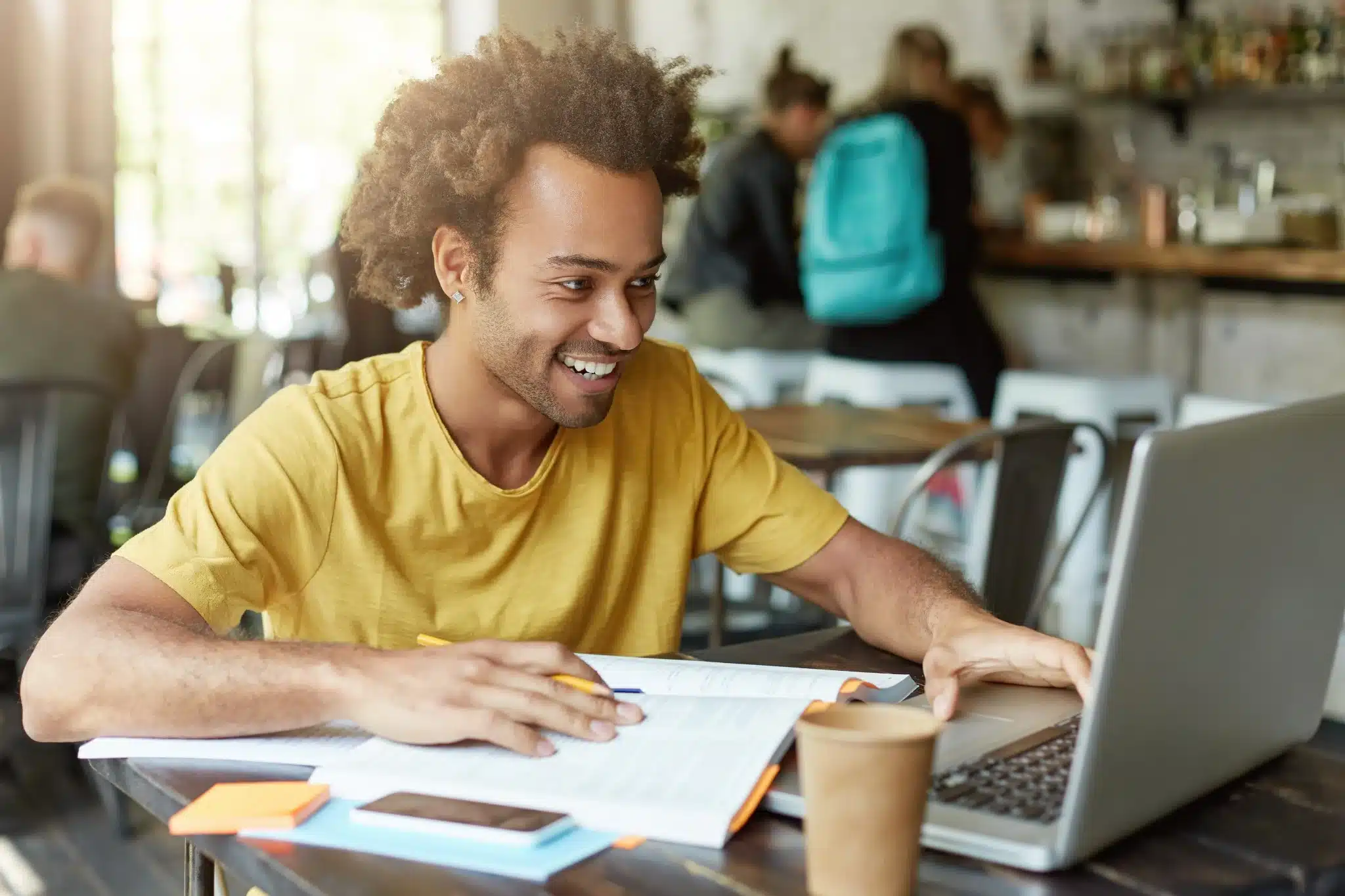 Estudante focado em seus estudos online na cafeteria Jovem sorrindo enquanto estuda com notebook e livro em cafeteria.