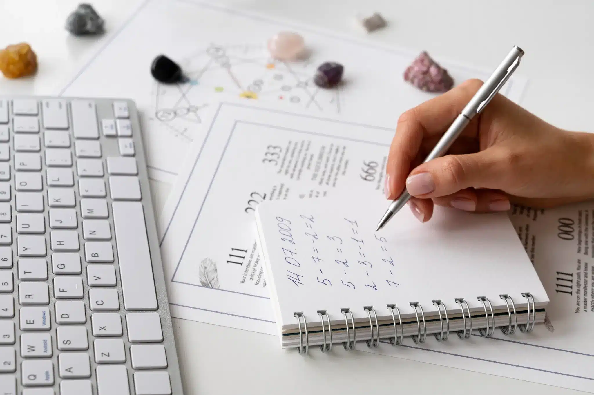 Numerologia Mão escrevendo anotações sobre numerologia em um caderno ao lado de cristais e um teclado.
