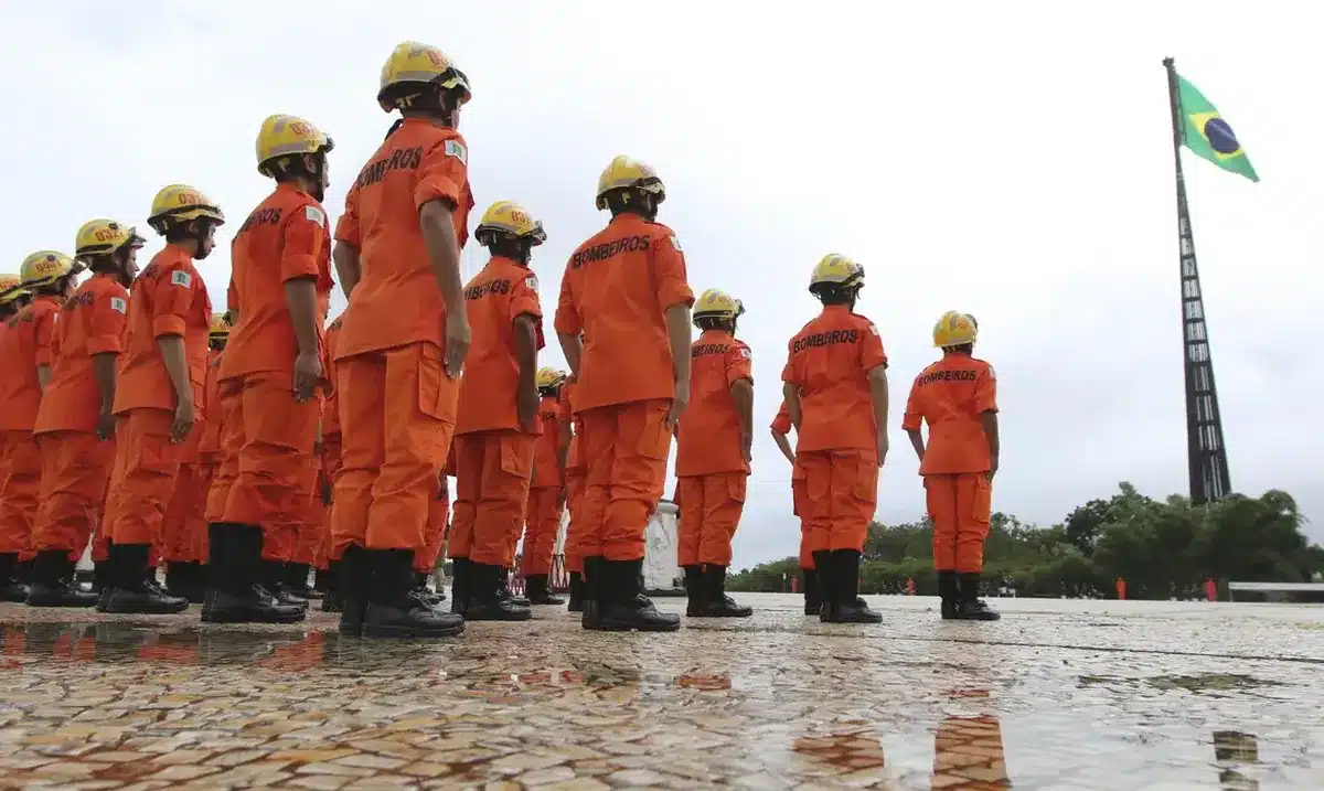 Concurso Bombeiro Bombeiros em formação durante cerimônia com bandeira do Brasil ao fundo.