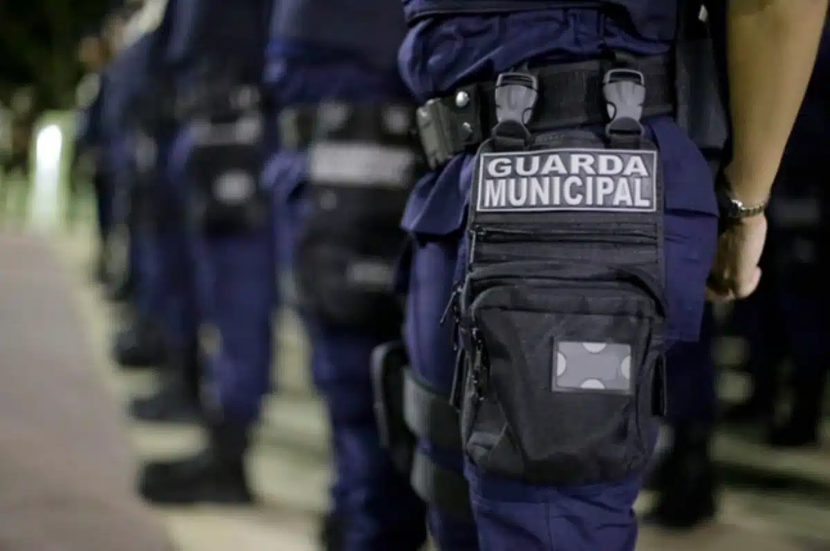 Conjunto de guardas municipais alinhados, focando no colete com identificação visível.