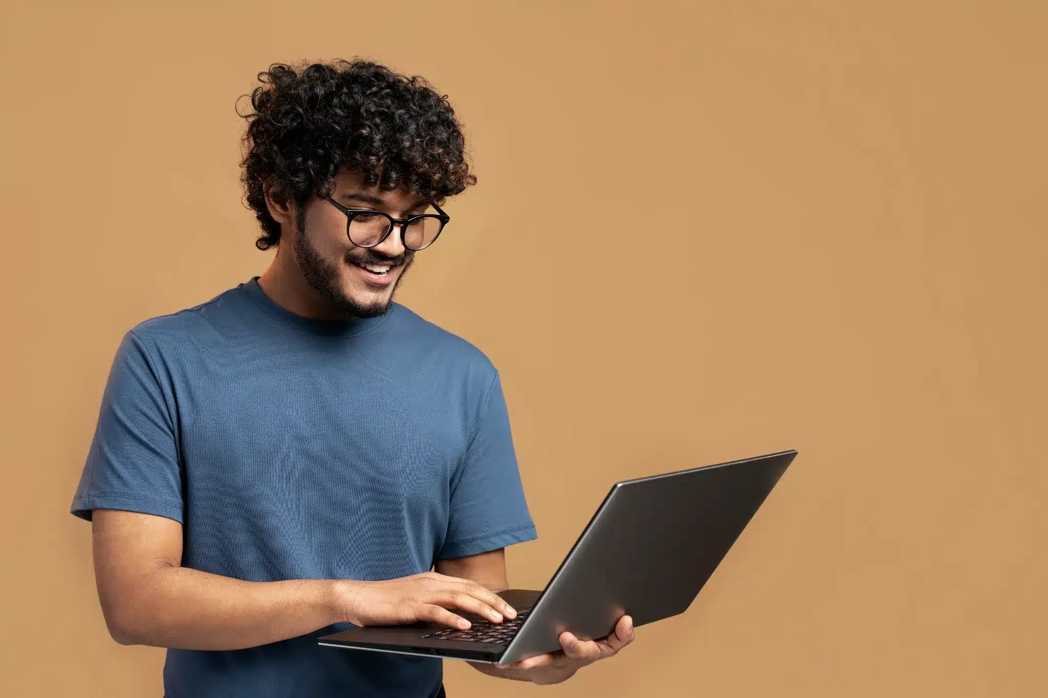 Homem sorridente usando óculos e camiseta azul, segurando e digitando em um notebook.