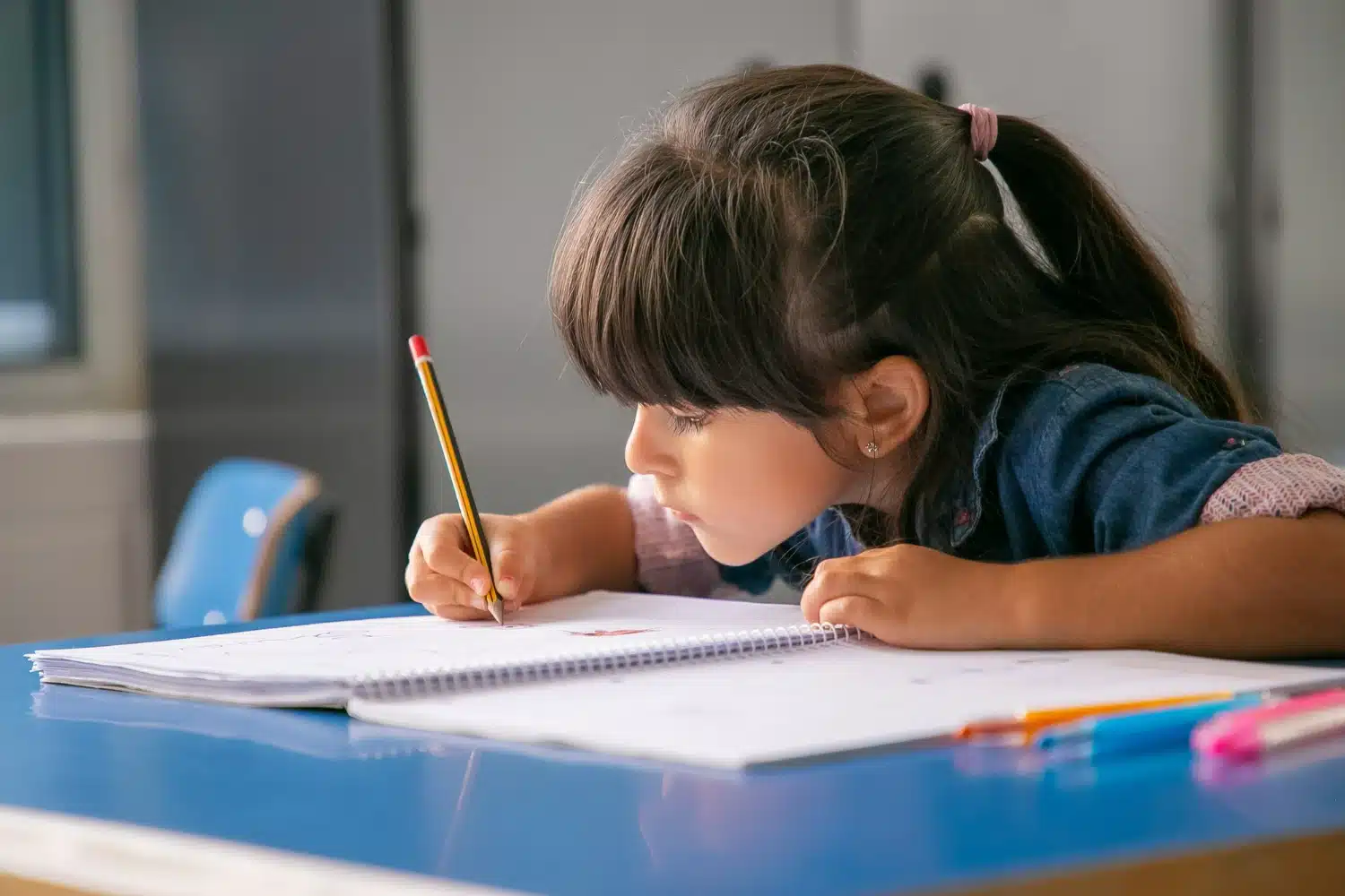 Criança focada estudando e escrevendo em caderno Menina concentrada escrevendo em caderno espiral, sentada à mesa azul em sala de aula.