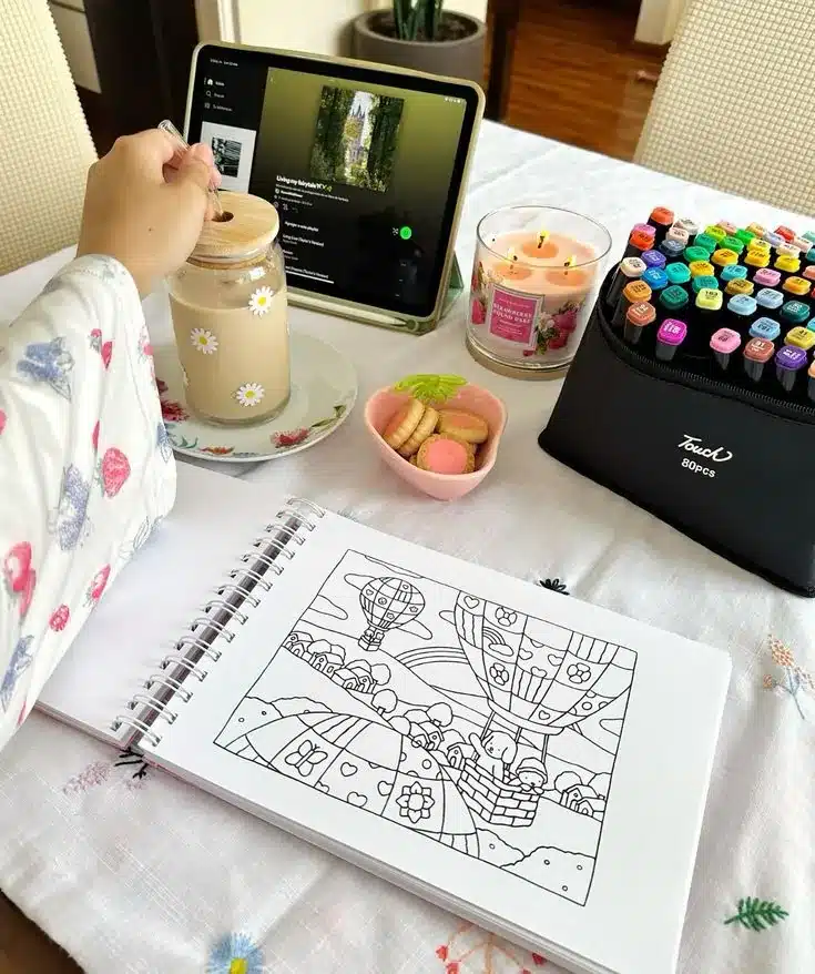 Momento de autocuidado com desenho, café e música Mesa organizada com café, biscoitos, vela aromática, tablet com música e livro de colorir.