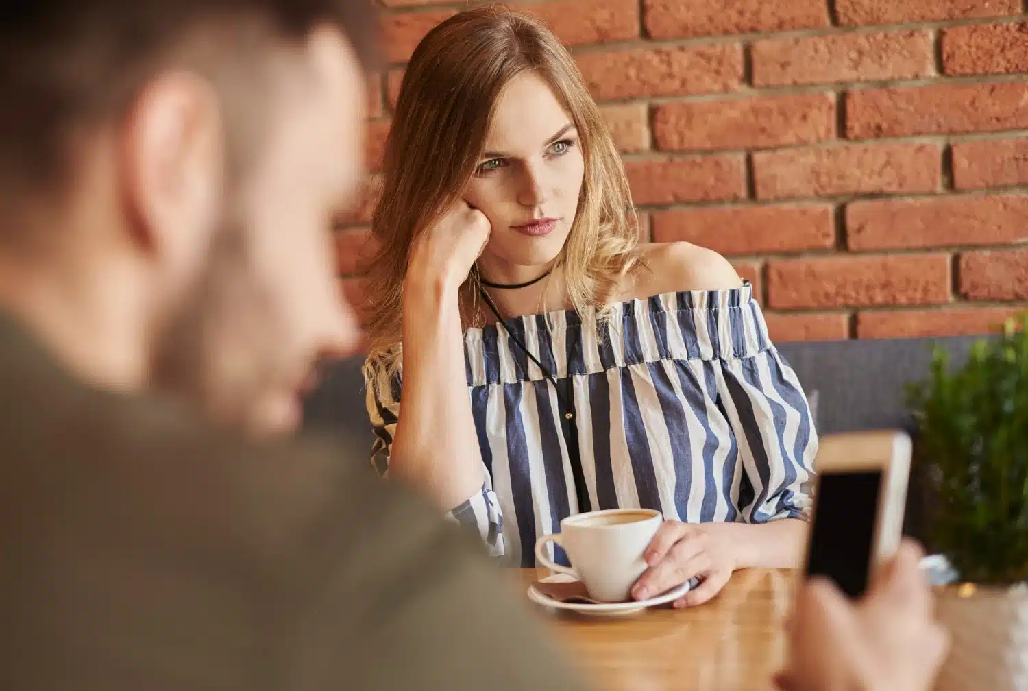 Mulher pensativa e séria segurando uma xícara de café enquanto homem ao fundo está distraído com o celular.