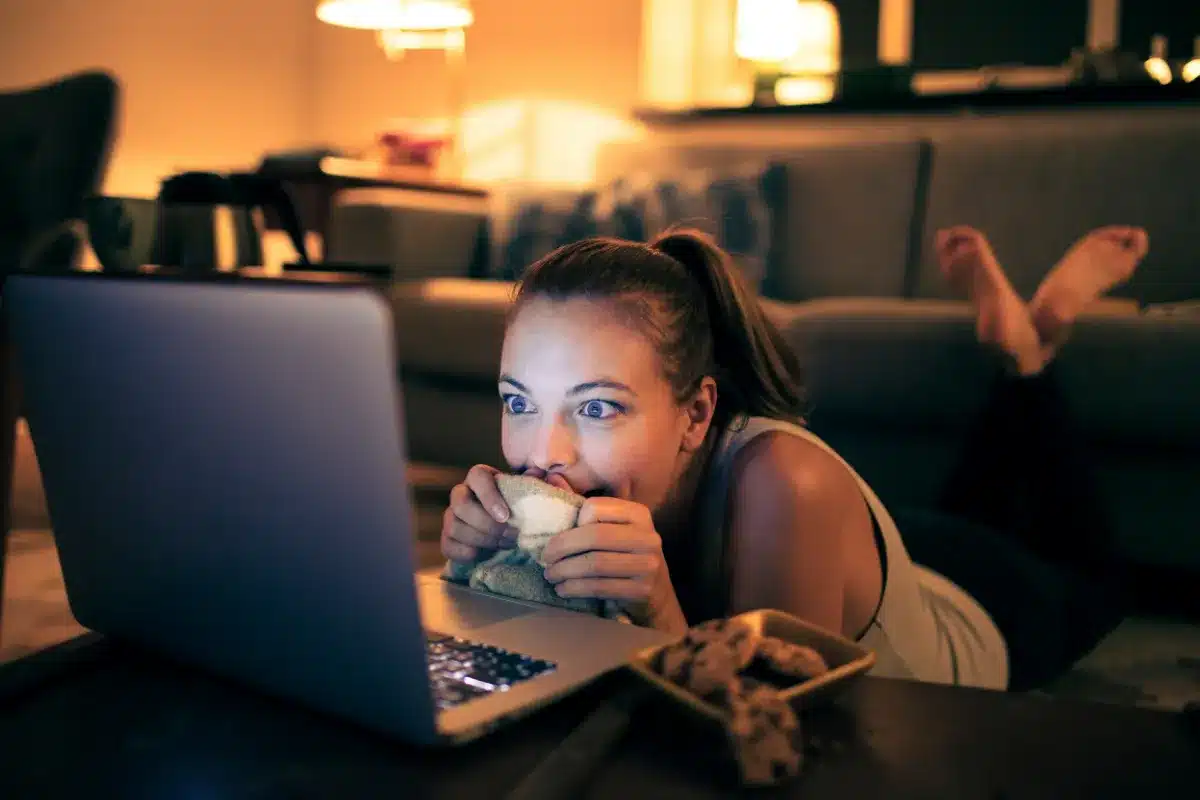 Mulher jovem deitada no chão, assistindo atentamente ao laptop com expressão animada, segurando uma manta.