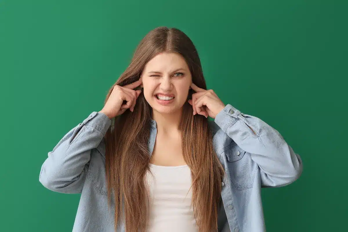 Mulher jovem tampando os ouvidos com os dedos e expressão de incômodo sobre fundo verde.