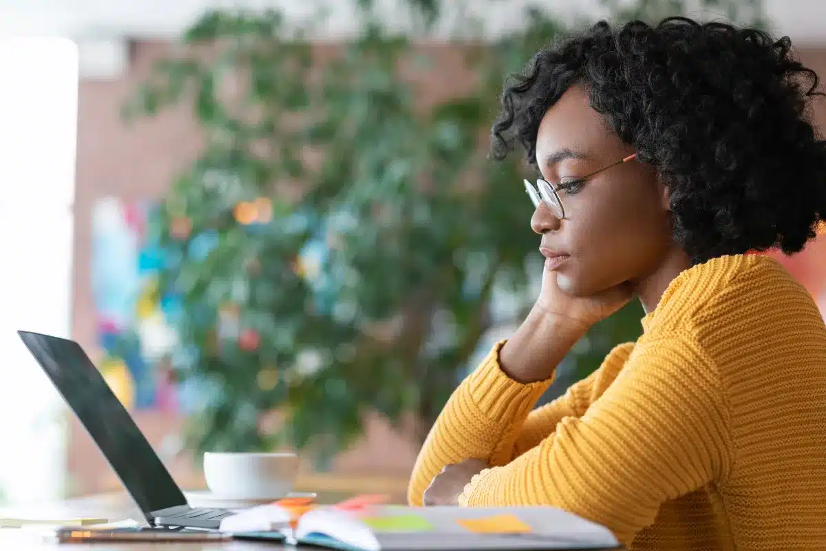 Mulher com cabelo cacheado e óculos, estudando em frente ao laptop com livros coloridos e uma xícara ao lado.