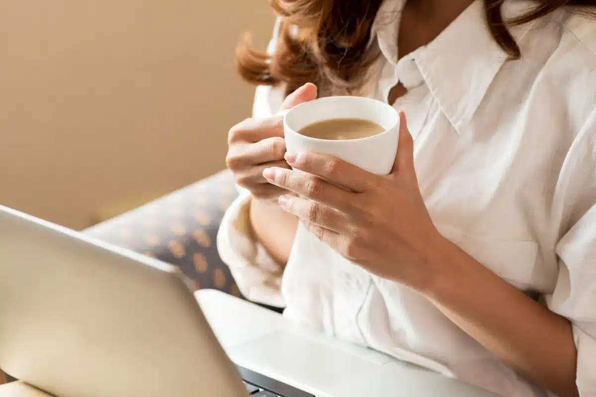 Café e foco: a pausa perfeita durante o trabalho Mulher segurando uma xícara de café com leite enquanto trabalha no notebook.