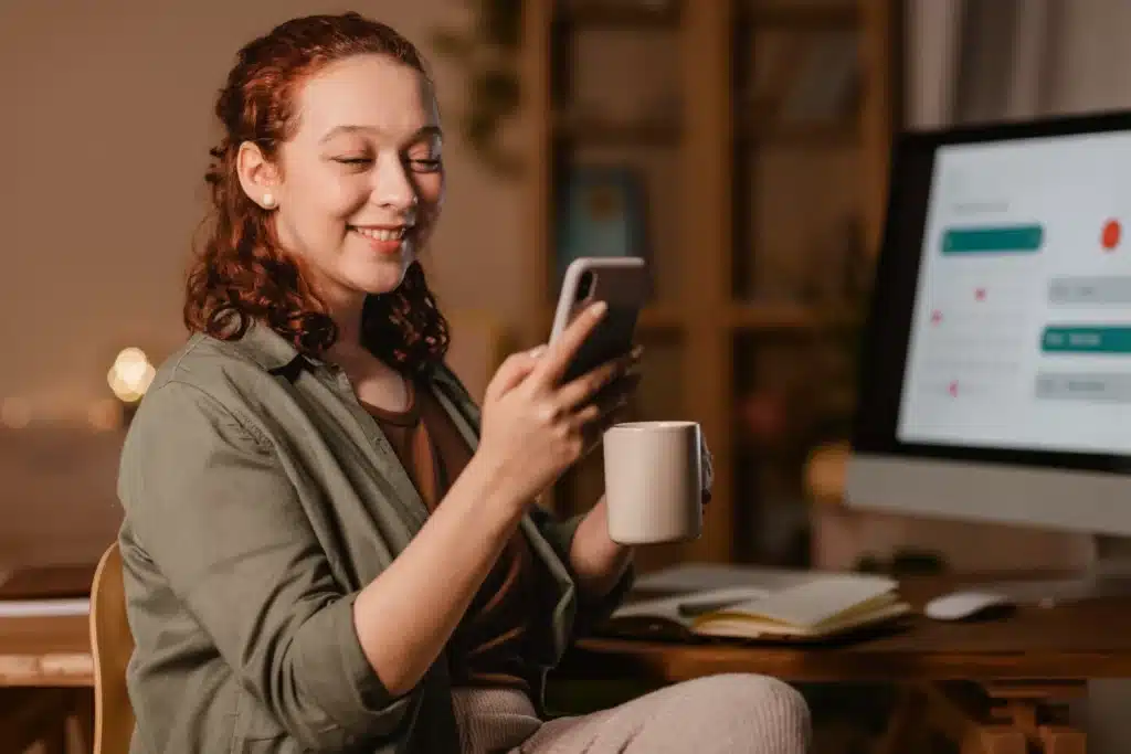 Preferência entre WhatsApp e Instagram revela traços pessoais Mulher sorrindo enquanto olha para o celular e segura uma caneca, em ambiente aconchegante com luz suave