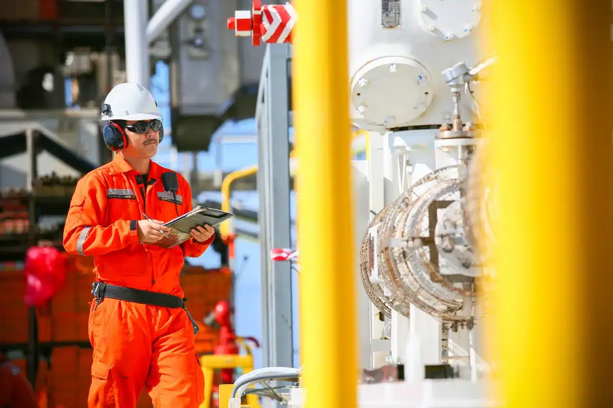 Engenheiro de Segurança de Processo com Tablet Engenheiro de segurança de processo, usando capacete e protetor auricular, fazendo anotações em um tablet em um ambiente industrial.